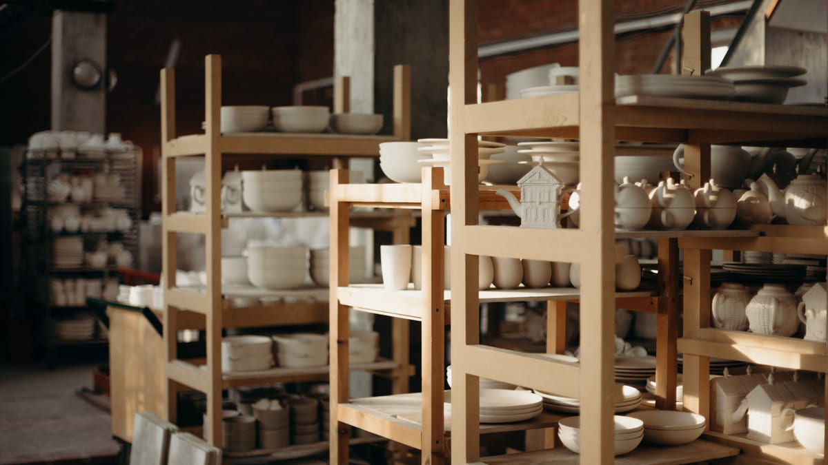 Rows of unfinished ceramic kitchenware on wooden shelves in a dim pottery workshop.
