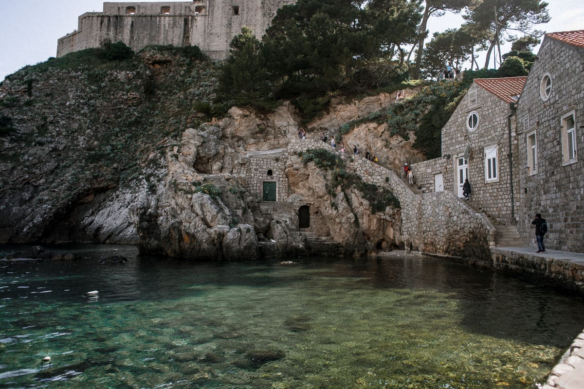 A rocky coastal inlet below a stone fortress and waterside walkway in Dubrovnik.