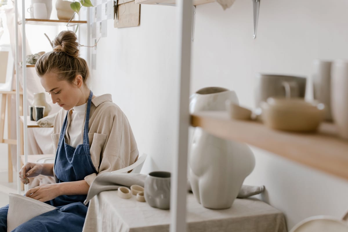 A potter at a sunlit bench, seen through shelves of pale ceramics.
