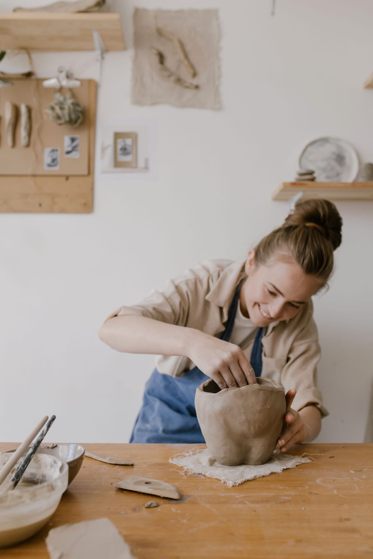 A maker in a blue apron shaping a pot beside tools and wall-mounted shelves.