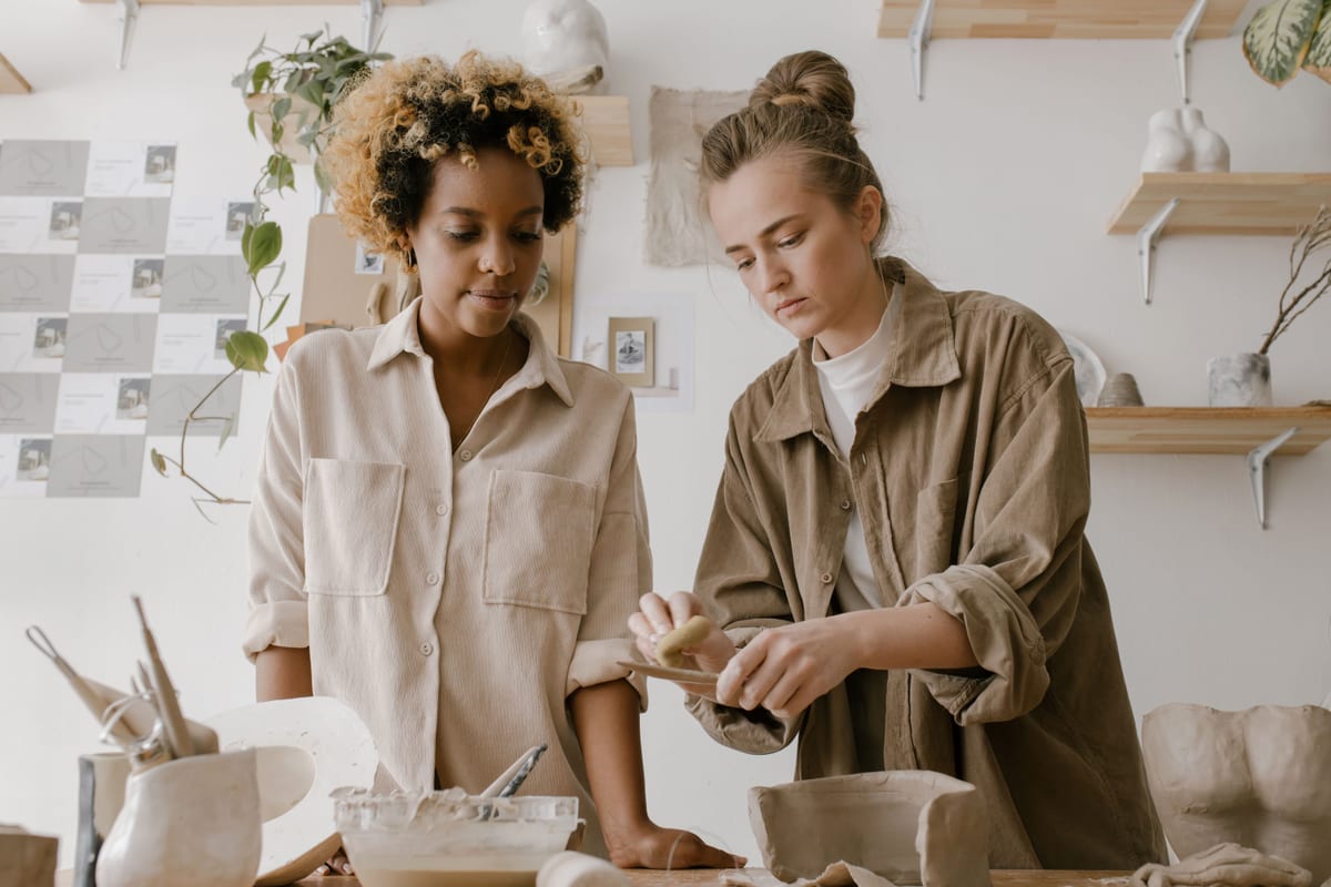 Two makers shaping clay at a wooden table in the white-walled ceramic studio.