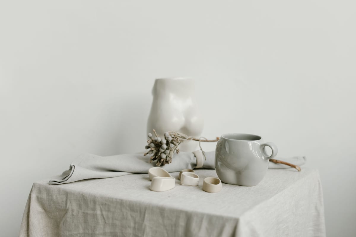 Pale ceramic vessels and small rings arranged on loosely draped linen.