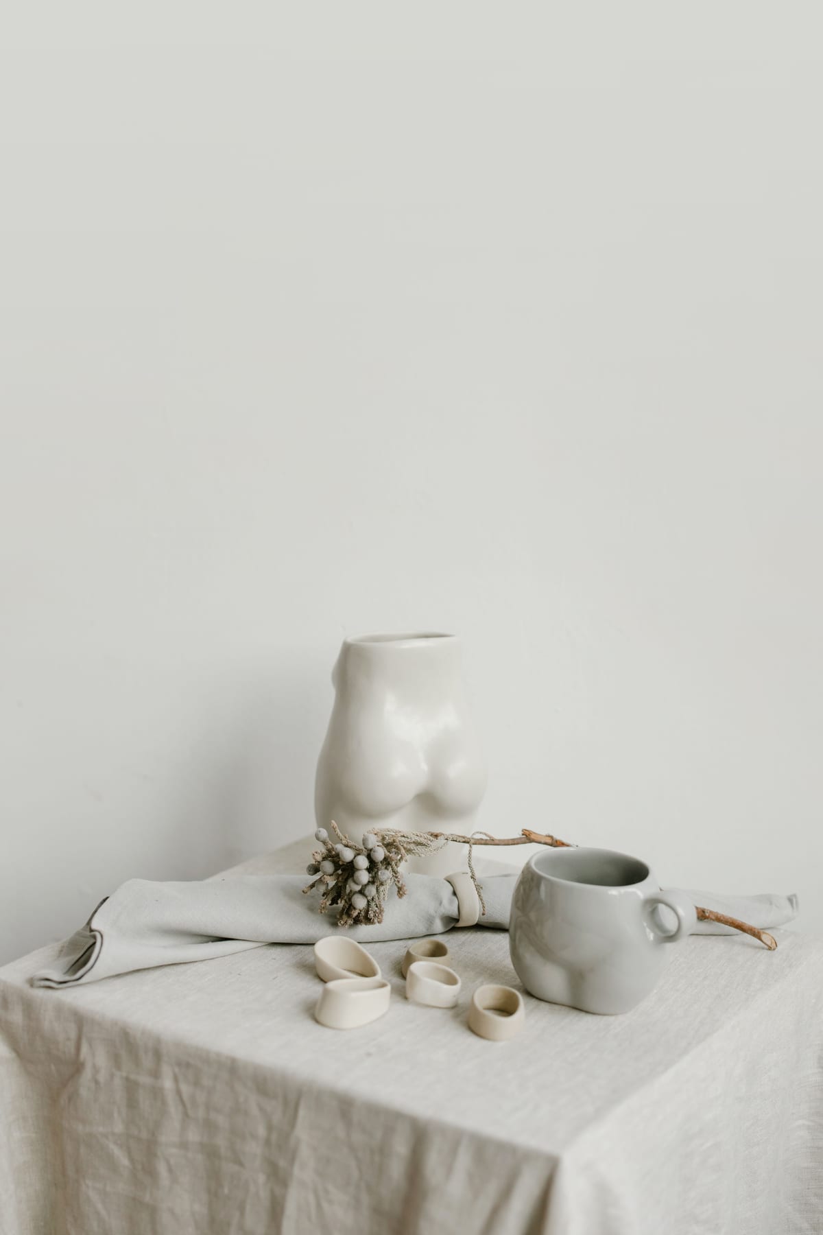 A tall sculptural white vase and a small cup on a linen-covered table against a plain wall.